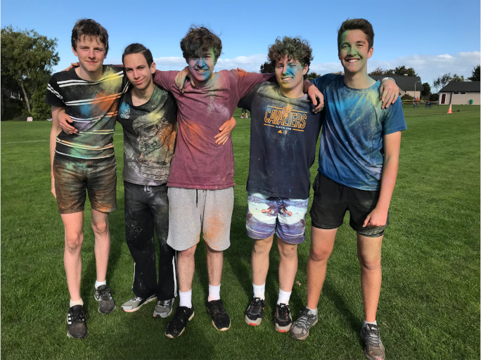 Five students standing with arms over each others shoulders, splattered with powerder paint